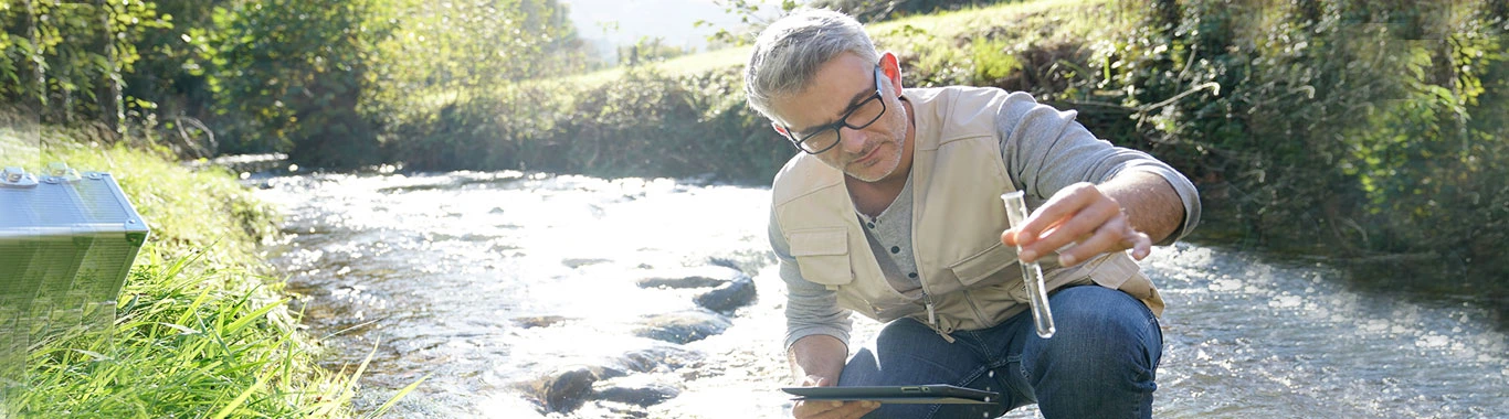 Man crouching by riverbank examining water quality or aquatic samples while wearing glasses and casual outdoor clothing