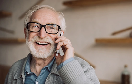 Senior man with gray hair and glasses smiling while talking on a phone, wearing a gray cardigan over a blue shirt in a home setting