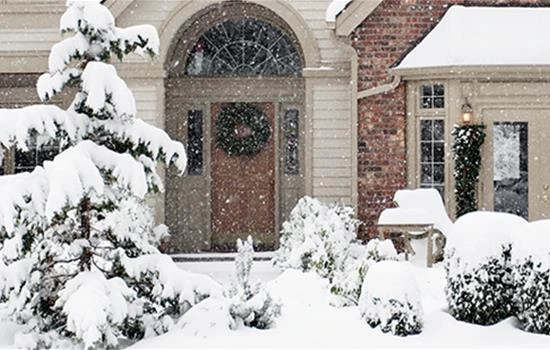 Snow-covered brick house entrance with wreath-adorned arched doorway, evergreen shrubs and trees blanketed in fresh white snow during winter