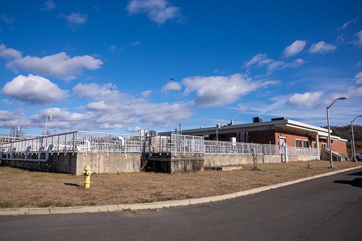 Exterior view of a wastewater treatment plant with a brick operations building and fenced treatment tanks.